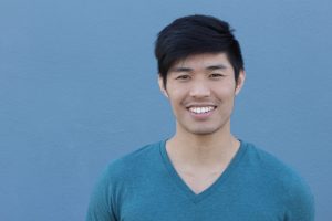 Young man with black hair and a blue V neck tshirt on smiling directly at the camera. He is standing in front of a plain blue background.