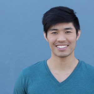 Young man with black hair and a blue V neck tshirt on smiling directly at the camera. He is standing in front of a plain blue background.