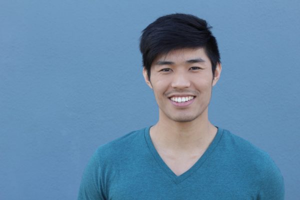 Young man with black hair and a blue V neck tshirt on smiling directly at the camera. He is standing in front of a plain blue background.
