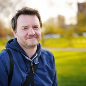 Man with brown hair and facial stubble, wearing a blue jacket and looking into the camera. He is standing in a park with grass behind him.