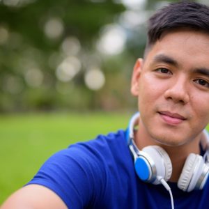 a young man with short dark hair is staring at the camera with a neutral expression. He is wearing a blue top and has white headphones around his neck.