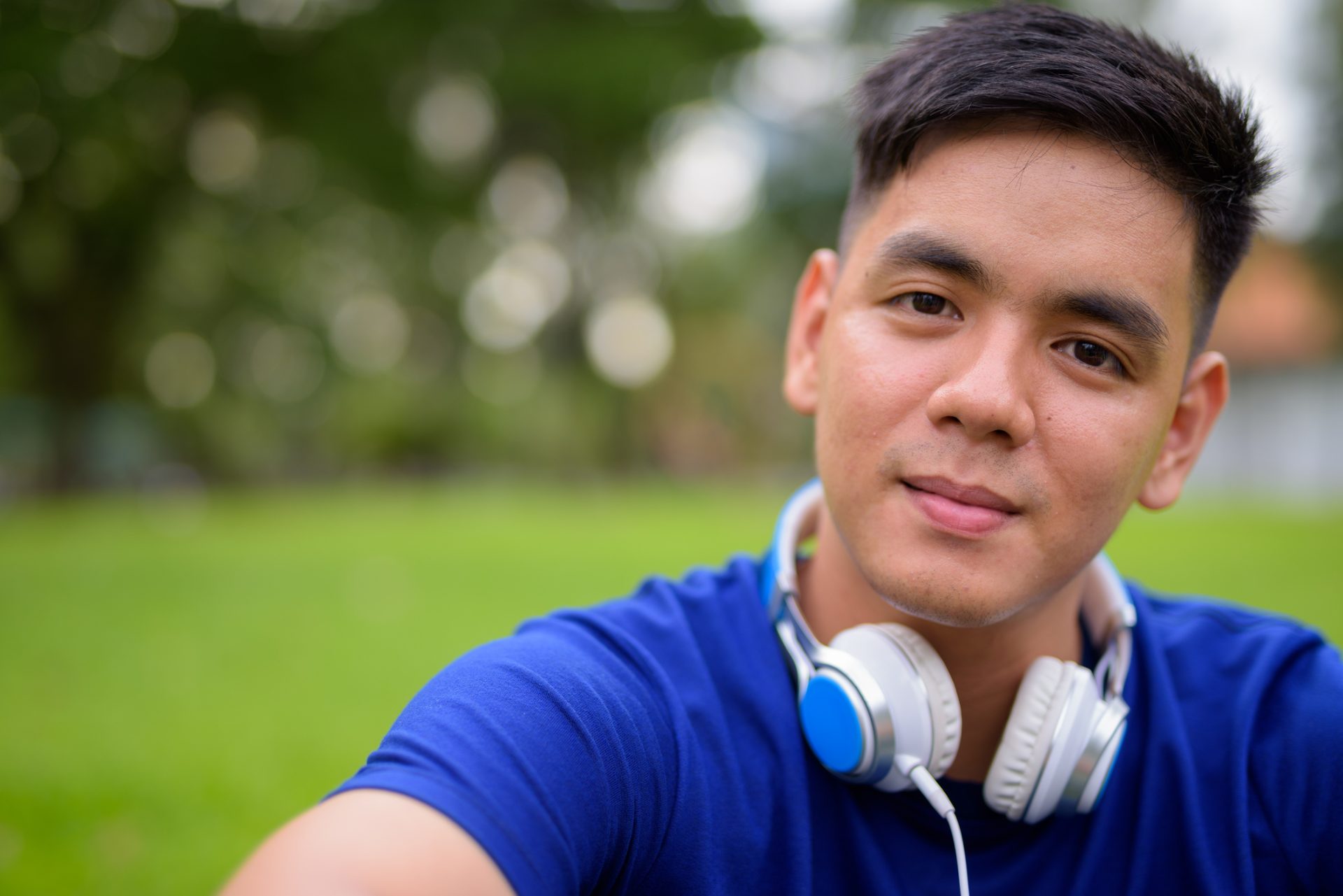 a young man with short dark hair is staring at the camera with a neutral expression. He is wearing a blue top and has white headphones around his neck.