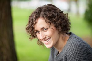 a young white women with long curly hair is looking to her left and smiling directly at the camera. The blurred background suggests she is in a green area, she is also wearing a grey jumper