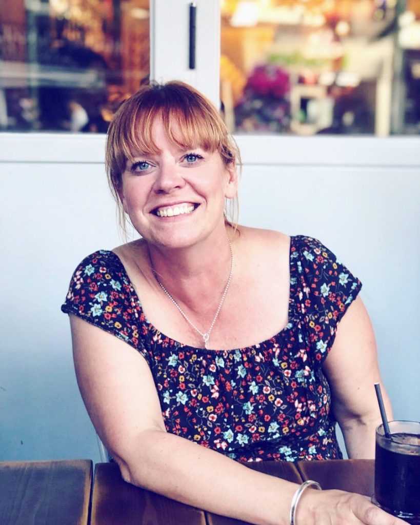 The photo shows our staff member Hils, who has blue eyes and ginger hair, she is sitting down inside holding a glass of coke and smiling at the camera