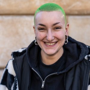 Young woman with bright green buzzcut hair. She has silver ear piercings and is wearing a black hoodie under a larger black and white coat. She is smiling widely, looking directly into the camera and is in front of a pale wall.