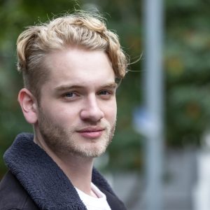 a young white man with blond curly hair and blond facial hair is smiling at the camera. He is wearing a white t shirt and a dark grey coat.