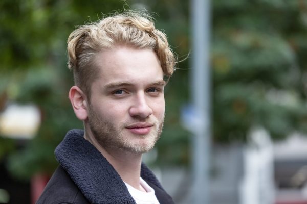 a young white man with blond curly hair and blond facial hair is smiling at the camera. He is wearing a white t shirt and a dark grey coat.