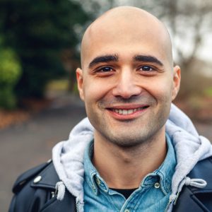 Young man who is bald, smiling directly at the camera. He is standing in a park with tree's behind him.