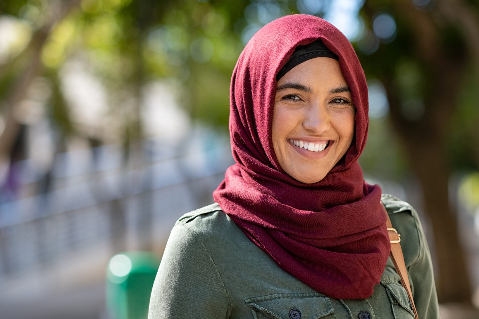 the photo shows a woman wearing a red hijab with brown eyes standing outside a building and smiling straight at the camera