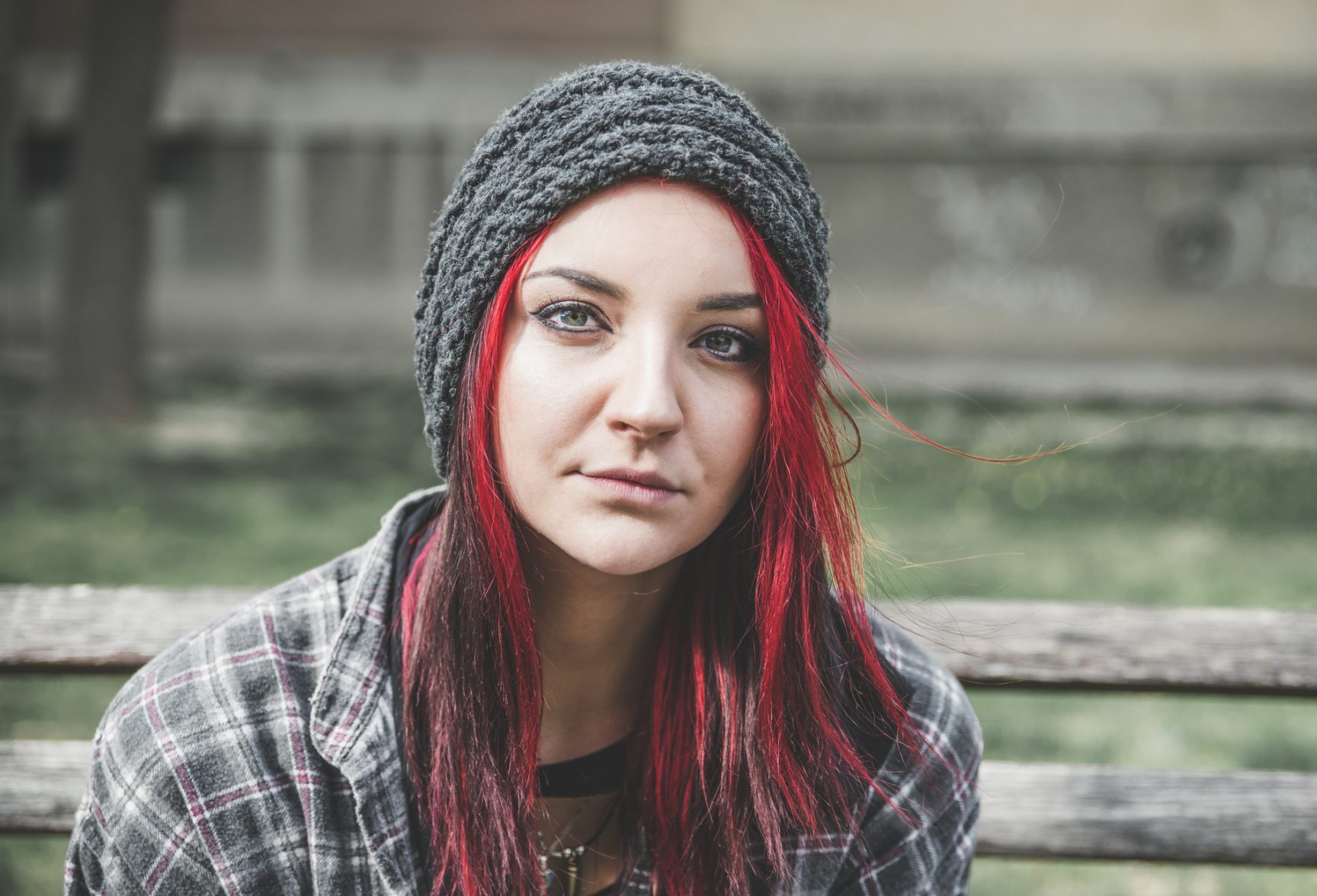 the photo shows a young girl sitting outside on a park bench, she has green eyes and long straight red hair, she is looking at the camera with a neutral expression