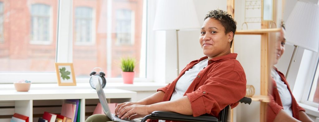 the photo shows a woman with short dark hair and dark eyes who is sitting inside on her wheelchair with her hands resting on her laptop keyboard