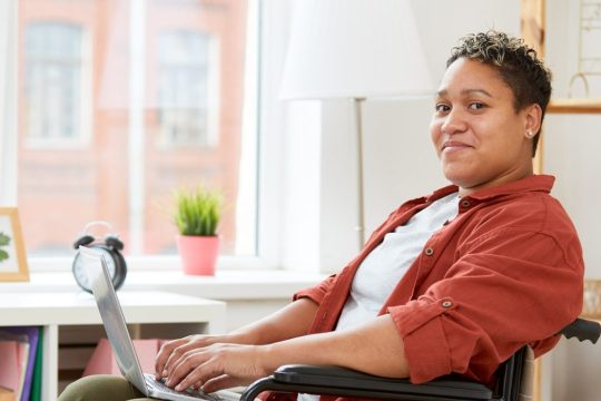 the photo shows a woman with short dark hair and dark eyes who is sitting inside on her wheelchair with her hands resting on her laptop keyboard