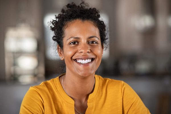 a women with short curly hair is smiling broadly at the camera, she is wearing a yellow jumper.
