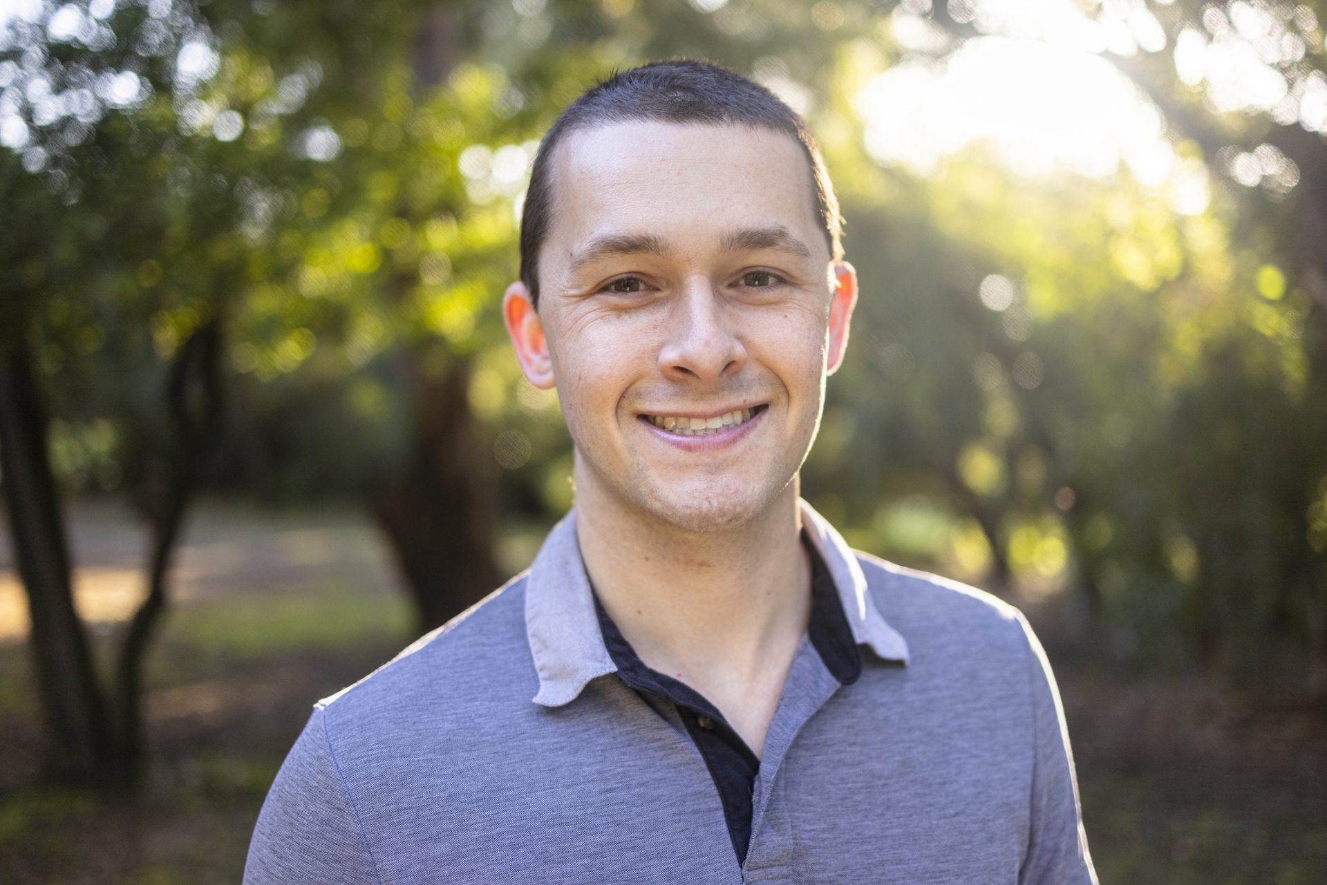 a young white male with short dark hair is smiling at the camera, he is wearing a light blue polo shirt