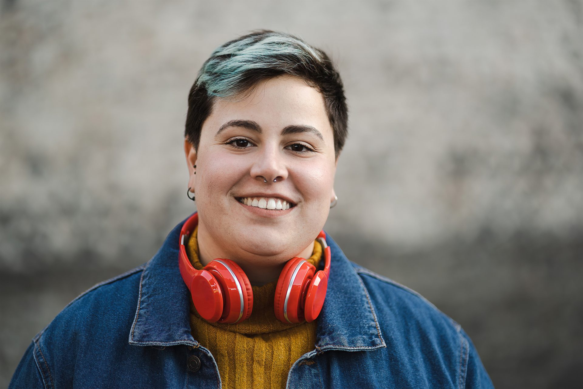 a young girl with short dark hair is smiling at the camera, her hair has green highlights and she has pierced ears and a nose piercing. She is wearing a jacket and has red headphones around her neck