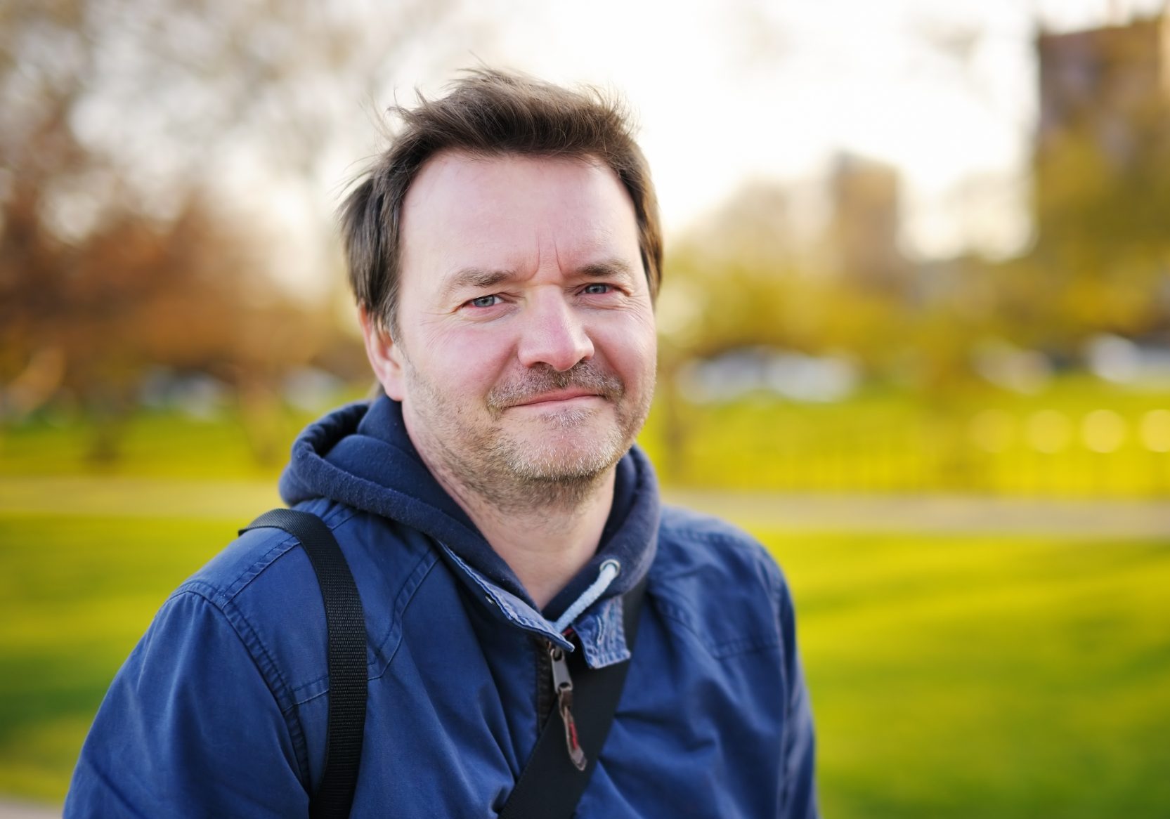 Man with brown hair and facial stubble, wearing a blue jacket and looking into the camera. He is standing in a park with grass behind him.