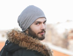 Young man with brown facial hair, a grey beanie hat and a black coat with fur along the hood. His body is facing away from the camera but he is looking over his right shoulder directly into the camera with a slight smirk.