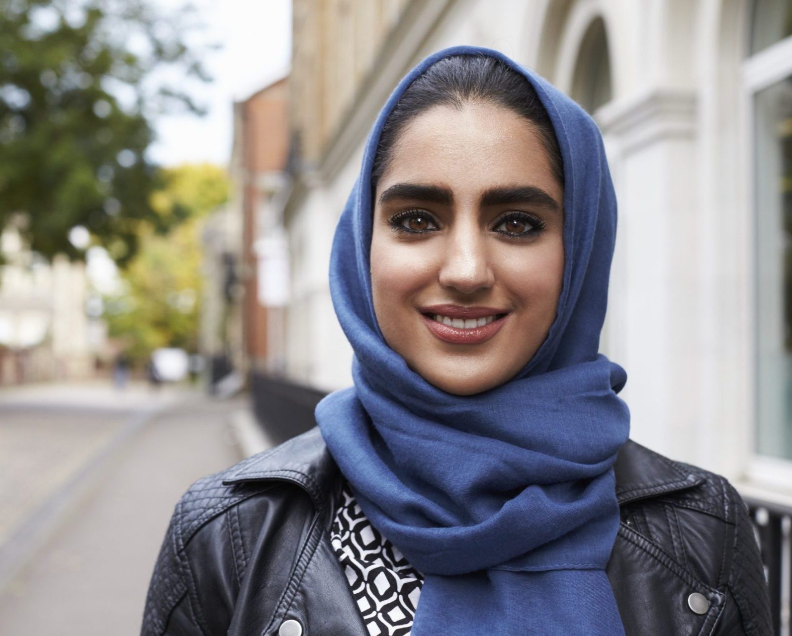 the photo shows a woman wearing a blue hijab with black hair and brown eyes standing outside a building and smiling straight at the camera