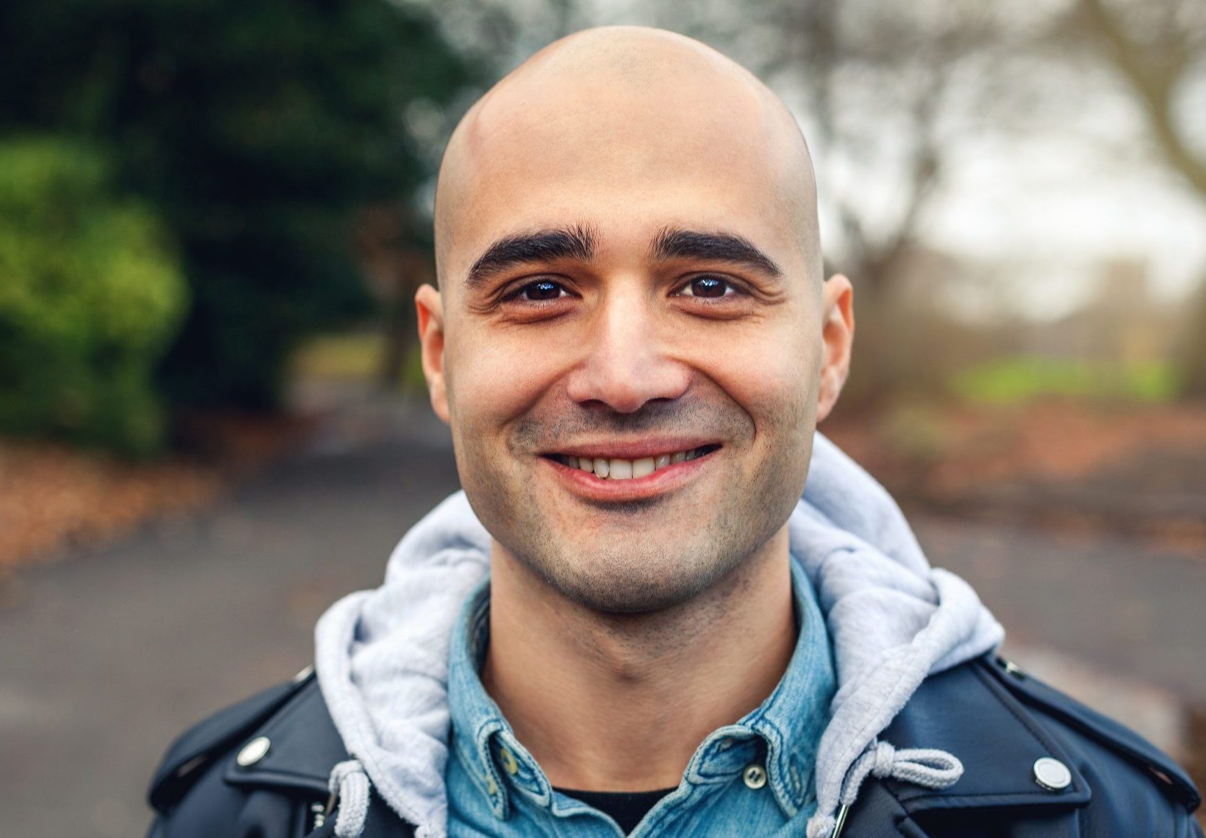 Young man who is bald, smiling directly at the camera. He is standing in a park with tree's behind him.