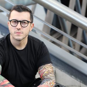 a young white man with short dark hair and glasses is sitting down on some stairs, he is wearing a black t shirt and looking at the camera with a neutral expression, he has tattoes on his arms.