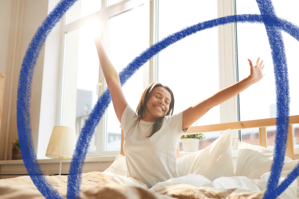 Young girl wearing a white tshirt with long brown hair, sat up in bed stretching her arms out. She is facing to the right of the camera and is smiling with her eyes shut. The bed has a wooden frame and white pillows and the windows show daylight.