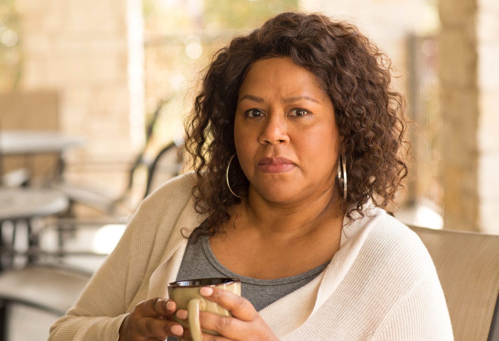 a women with shoulder length curly hair is smiling at the camera, she is holding a cup of coffee.