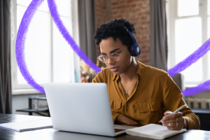 a young girl with glasses and dark, curly hair is sitting down at a table and looking at her laptop while writing in a notebook with her pen. She is wearing an orange shirt and has headphones around her ears.