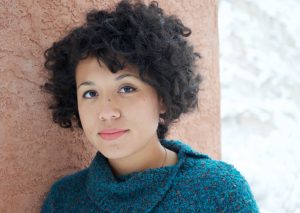 a women with short curly hair is smiling at the camera, she is leaning against a wall.
