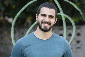 Young man with short black hair and facial hair, wearing a blue top and smiling directly in the camera.