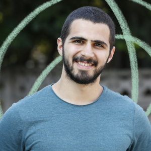 Young man with short black hair and facial hair, wearing a blue top and smiling directly in the camera.