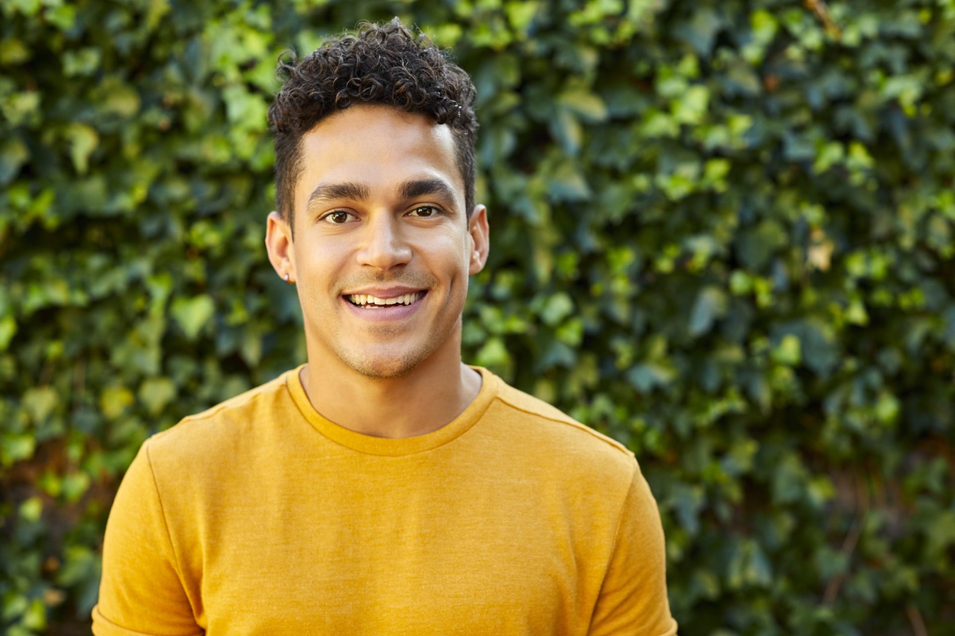 the photo shows a young man with short dark curly hair and eyes standing outside and smiling at the camera