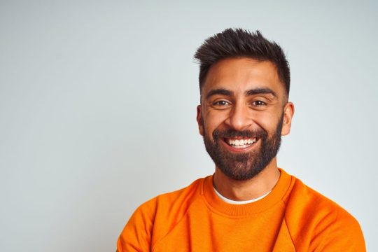 a young man with dark hair, eyes and a beard is standing inside and smiling at the camera with his arms folded