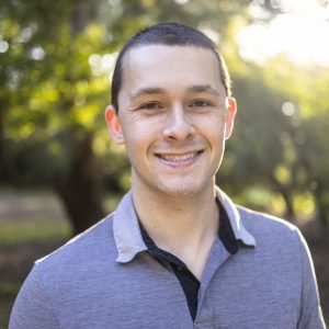 a young white male with short dark hair is smiling at the camera, he is wearing a light blue polo shirt