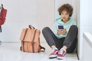A young boy sat on the floor in a school hallway, looking at his phone.