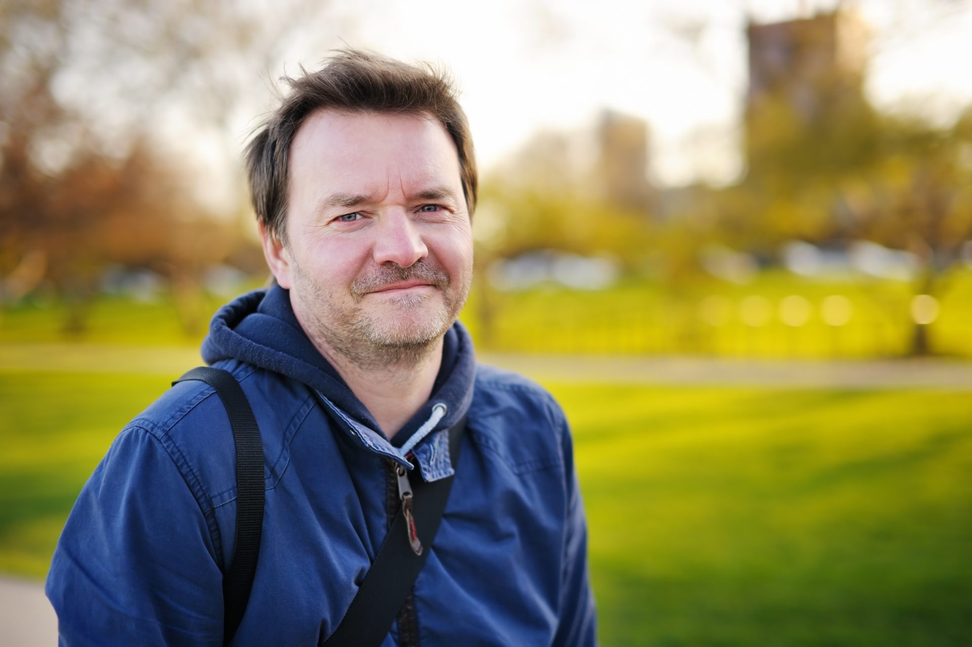 Man with brown hair and facial stubble, wearing a blue jacket and looking into the camera. He is standing in a park with grass behind him.