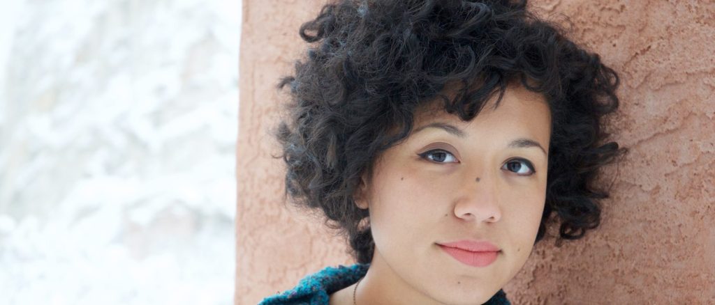 a women with short curly hair is smiling at the camera, she is leaning against a wall.