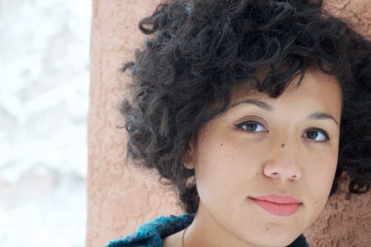a women with short curly hair is smiling at the camera, she is leaning against a wall.