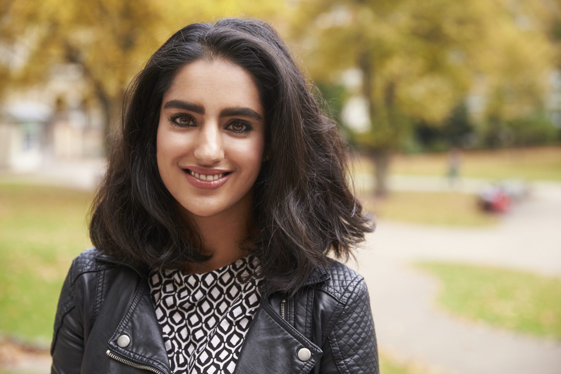 a women with dark, shoulder length hair is smiling broadly at the camera, she is wearing a patterned top and a black leather coat.