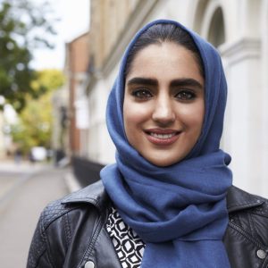Young woman in blue hijab smiling directly into the camera. She is standing on the pavement of a residential street.