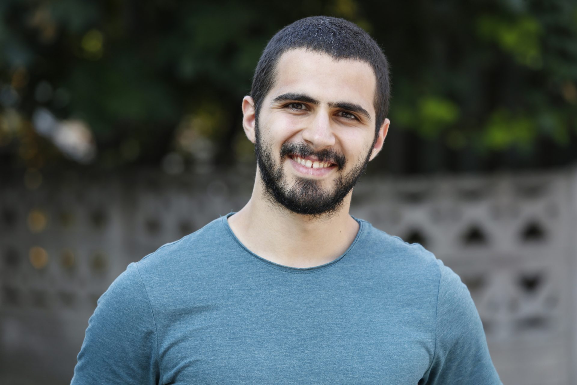 the photo shows a man with short black hair and brown eyes standing outside and smiling straight at the camera