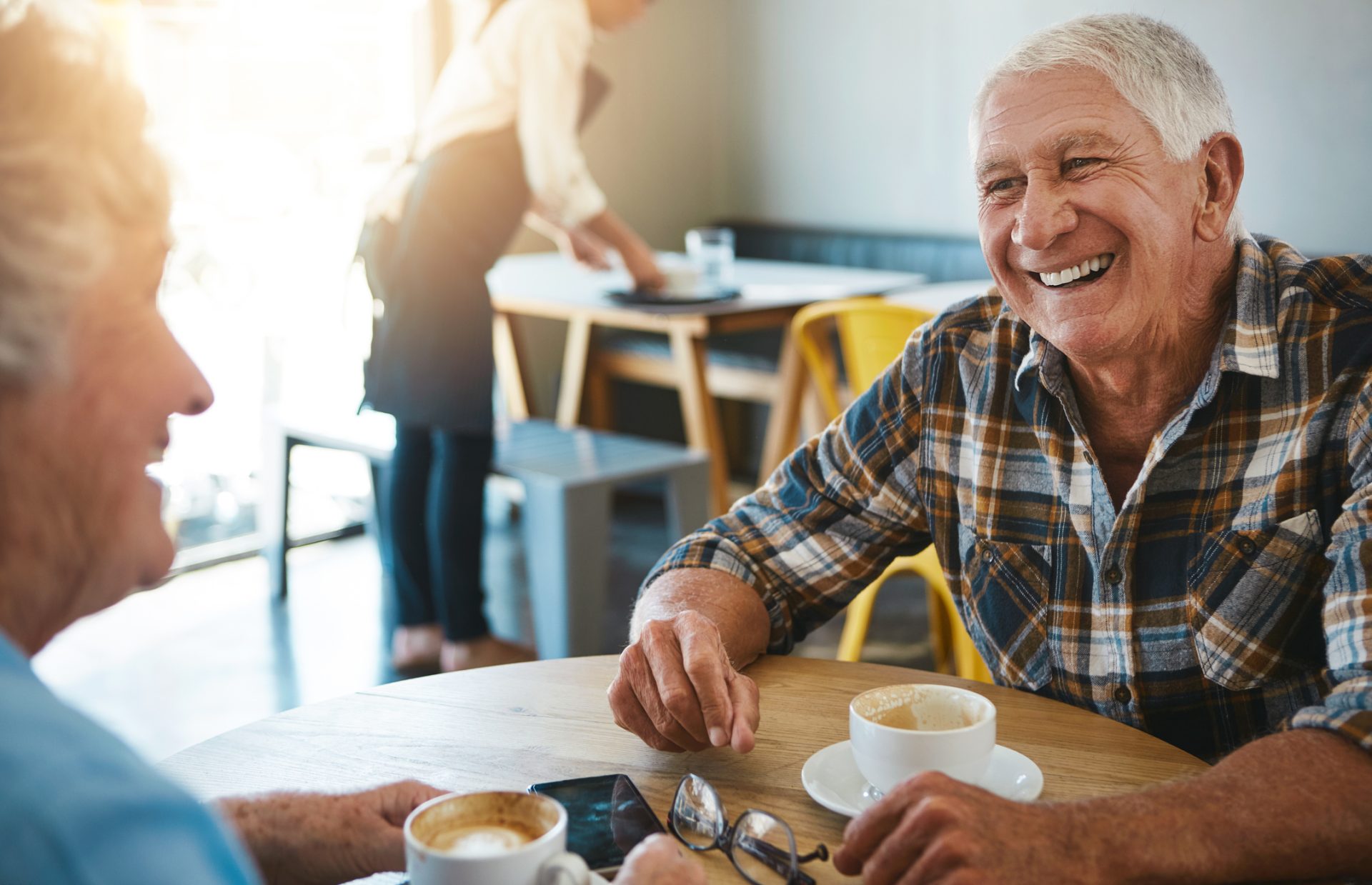 An older man, smiling and having a cup of coffee with someone.