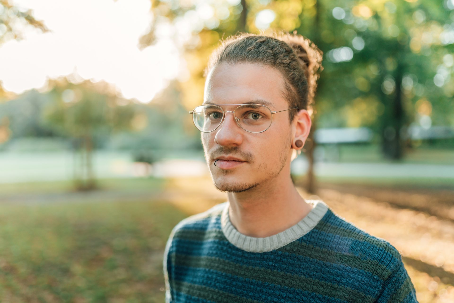 the photo shows a man with ponytail hair and glasses standing outside in a park, looking at the camera with a serious expression