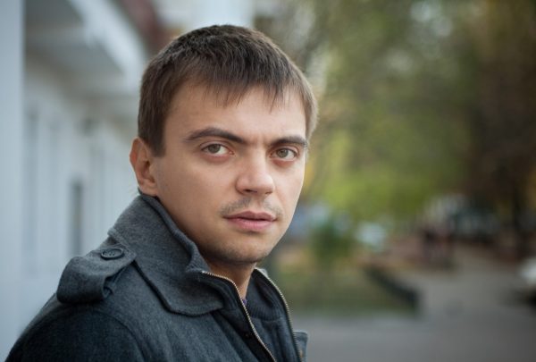 a man is looking to his right and staring directly into the camera with a neutral expression. He has short, straight hair and is wearing a grey coat.