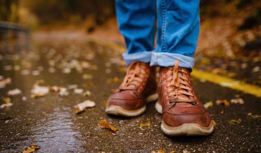 the photo shows a pair of smart brown shoes and blue jeans standing outside on a concrete path with leaves