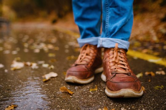 the photo shows a pair of smart brown shoes and blue jeans standing outside on a concrete path with leaves